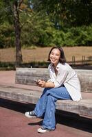 Happy smiling asian girl sitting on bench in park and browsing on social media, holding smartphone, using mobile phone app photo
