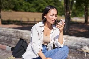 Asian girl frowning, looking concerned at her mobile phone, reading sad message, bad news on smartphone app, sitting on bench in park worrying photo