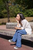 Portrait of asian girl sitting with smartphone feeling sad, looking gloomy and frustrated, waiting for a call outdoors in park photo