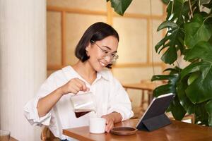 Lanza libre y remoto trabajadores sonriente joven mujer torrencial café en un taza, sentado en café y mirando a digital tableta foto