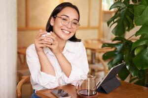 sonriente asiático niña en anteojos, mujer trabajando en remoto, Bebiendo café y utilizando tableta foto