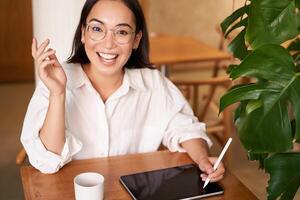 Young asian creative girl, sitting with digital tablet, drawing with graphic pen and smiling, doodling, drinking coffee in cafe photo