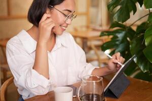 Portrait of beautiful young woman sitting in cafe, drinking coffee and drawing diagram on tablet with graphic pen, smiling pleased photo