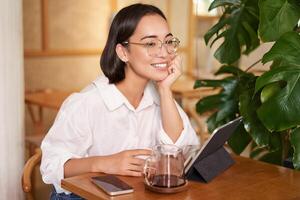 sonriente asiático niña en anteojos, mujer trabajando en remoto, Bebiendo café y utilizando tableta foto