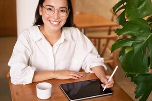 Smiling young female creator, creative girl drawing on digital tablet with graphic pen, drinking coffee in cafe photo