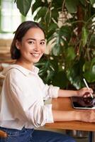 vertical Disparo de joven asiático mujer con digital tableta y gráfico bolígrafo, dibujo, rascarse mientras sentado en café con taza de café foto