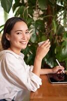 vertical Disparo de joven asiático mujer con digital tableta y gráfico bolígrafo, dibujo, rascarse mientras sentado en café con taza de café foto