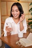 Portrait of modern girl sits with coffee in cafe, smiling while looking at smartphone, reading book in restaurant and using mobile phone app photo