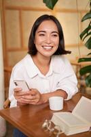 Portrait of modern girl sits with coffee in cafe, smiling while looking at smartphone, reading book in restaurant and using mobile phone app photo