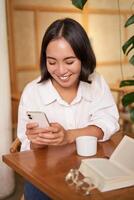 Portrait of modern girl sits with coffee in cafe, smiling while looking at smartphone, reading book in restaurant and using mobile phone app photo