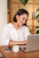 sonriente hembra gerente, persona de libre dedicación o estudiante sentado con ordenador portátil en café y laboral, mecanografía en computadora foto