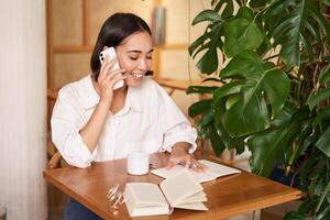 trabajando mujer responder teléfono llamada en cafetería, escritura abajo, haciendo notas mientras teniendo conversacion en teléfono foto