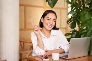 Stylish young entrepreneur, girl with laptop sitting in cafe, pointing finger at advertisement banner on upper left corner photo