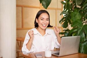 entusiasta joven hembra gerente, mujer de negocios con computadora portátil, celebrando éxito, triunfando, sentado con ordenador portátil en café foto