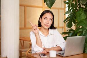 Thoughtful girl pointing at upper left corner, sitting in cafe with laptop and working, showing copy space banner photo