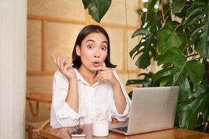 Girl with surprised face, pitching an idea, pointing left, sitting with laptop in cafe and drinking coffee photo