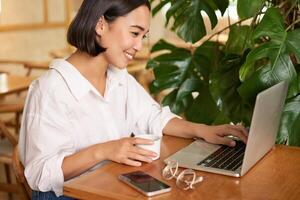 Student sitting in cafe with cup of coffee. Young asian woman working on laptop in restaurant, sitting with computer and smartphone photo