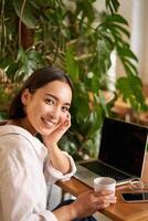 Trendy young woman sitting in cafe and smiling at camera, drinking coffee and using laptop, working remotely, studying or browsing internet photo