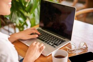 Cropped shot of laptop screen and female hands typing, cafe interior and cup of coffee with glasses on table photo