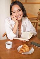 vertical Disparo de contento joven asiático mujer disfruta lectura, sentado con libro en cafetería, Bebiendo café y comiendo cuerno foto