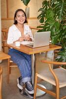 Vertical shot of young asian girl sits in cafe with laptop and smartphone, relaxing and surfing the net, working remotely photo