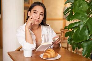 Young woman feels uneasy, reads sad book and sits in cafe with cup of coffee, looks around with sulking unhappy face photo