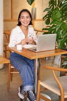 Vertical shot of young asian girl sits in cafe with laptop and smartphone, relaxing and surfing the net, working remotely photo