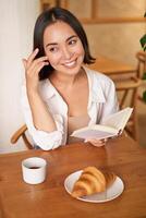 vertical Disparo de contento joven asiático mujer disfruta lectura, sentado con libro en cafetería, Bebiendo café y comiendo cuerno foto