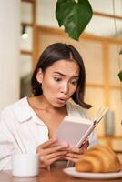 Vertical shot of asian girl looking shocked and worried at book, reading with interest, gasping from story plot, sitting in cafe with croissant and coffee photo