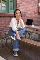 Vertical shot of trendy asian girl sitting in cafe alone with laptop, working or studying, browsing on computer photo