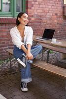 Vertical shot of trendy asian girl sitting in cafe alone with laptop, working or studying, browsing on computer photo