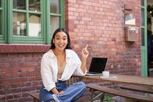 stylish modern asian girl with laptop, sitting in cafe, looking amazed and pointing at upper right corner banner, showing info advertisement photo