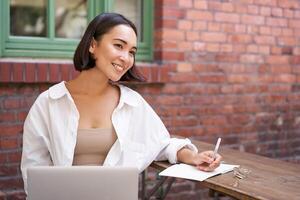 Beautiful young female model sitting with laptop in outdoor cafe, taking notes, writing in her diary or working on project, doing homework photo