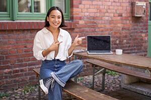 Stylish young asian woman sitting in cafe with laptop, outdoor table, pointing finger at advertisement, showing banner copy space photo