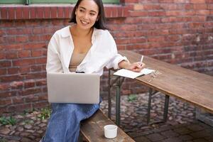 Working woman sitting outdoors in cafe, looking at laptop and making notes in notebook, writing down while watching smth on computer photo
