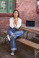 Full size portrait of asian girl sits with laptop, looks amazed and surprised and peeks at screen. Concept of people and urban lifestyle photo