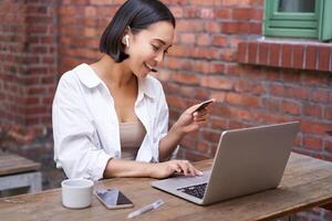 sonriente asiático mujer sentado con computadora portátil, pago por crédito tarjeta para en línea compras, enviando su banco cuenta detalles, sentado en café foto