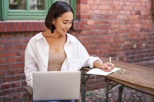 Working woman sitting outdoors in cafe, looking at laptop and making notes in notebook, writing down while watching smth on computer photo