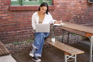 Working woman sitting outdoors in cafe, looking at laptop and making notes in notebook, writing down while watching smth on computer photo