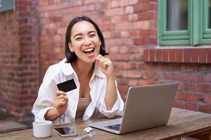 contento joven asiático mujer sentado cerca computadora portátil, participación crédito tarjeta, pago facturas, compras en línea sin contacto, sonriente a cámara foto