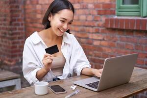 sonriente asiático mujer sentado con computadora portátil, pago por crédito tarjeta para en línea compras, enviando su banco cuenta detalles, sentado en café foto