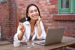 Enthusiastic asian girl sitting with credit card and laptop, paying contactless, shopping online via computer photo