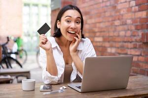 Portrait of young stylish girl sits near laptop, shows credit card and smiles, pays bills, shops online. photo