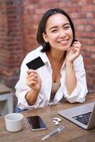 Beautiful young asian woman with credit card, sitting near laptop and smiling, paying bills, shops online, order smth on computer photo