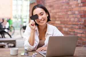 Happy young asian female, sitting with laptop and showing credit card, paying bills online, shopping in internet, sitting in cafe with coffee photo