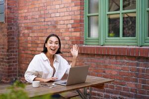 simpático asiático mujer sentado con computadora portátil, ondulación a tú, diciendo Hola, Hola gesto, saludo usted mientras trabajando con computadora foto