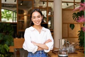 Young asian woman, cafe owner or manager, standing confident with arms crossed on hands, smiling and looking at camera photo