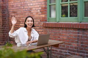 simpático asiático mujer sentado con computadora portátil, ondulación a tú, diciendo Hola, Hola gesto, saludo usted mientras trabajando con computadora foto