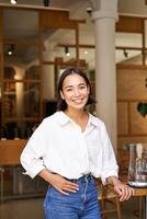 Smiling asian manager, confident woman standing near restaurant entrance, cafe owner welcomes guests photo