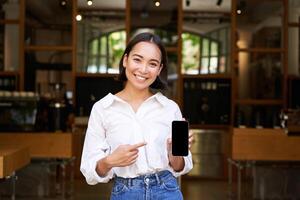 Happy asian woman, cafe manager standing in front of restaurant, pointing finger at smartphone app, showing mobile screen, recommending application photo